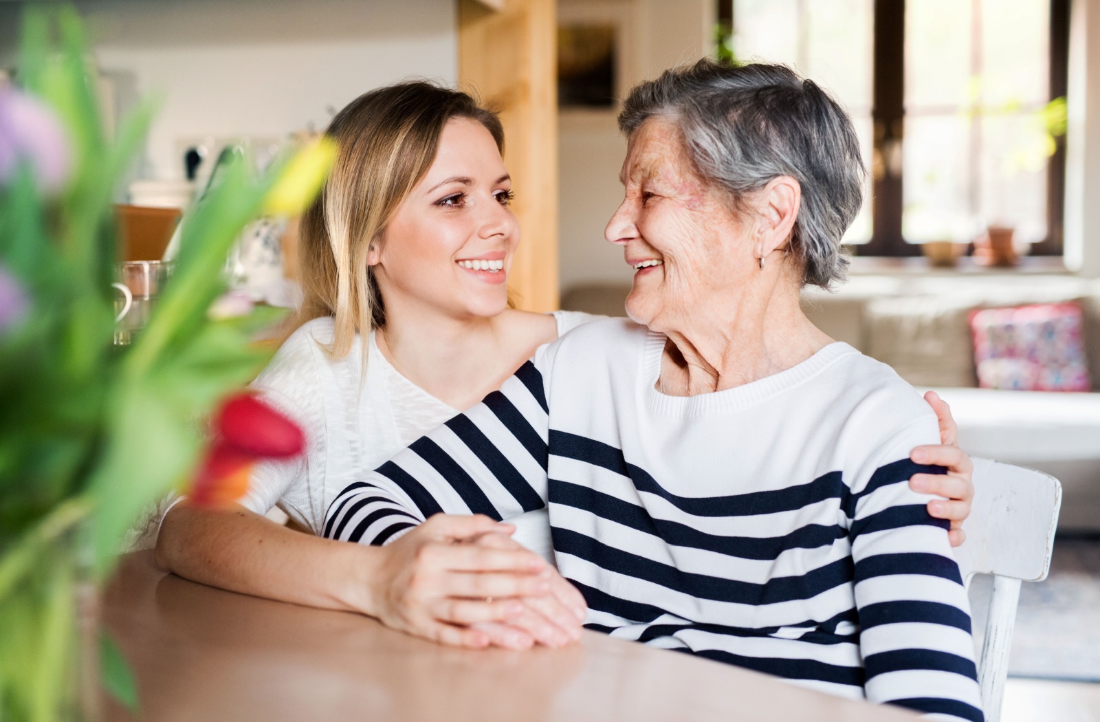 An older adult woman being embraced by her daughter as they smile at each other.