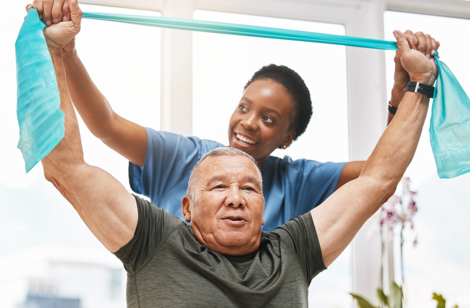 A staff member helping an elderly man stretch his arms over his head with the help of a stretch band.