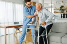 A caregiver helping a senior man use his walker to get off the couch.