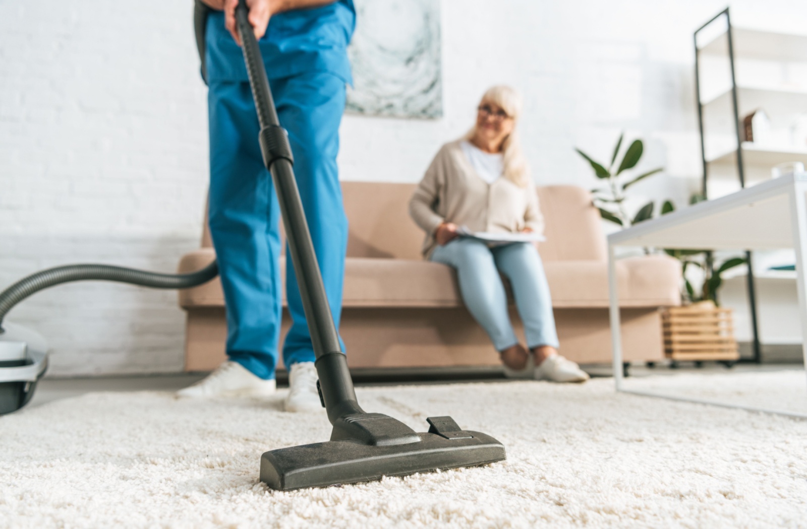 A close-up image of a caregiver vacuuming a white carpet while a senior woman sits on the couch smiling.