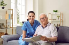 A caregiver and a senior man in assisted living smiling at the camera while sitting on the couch looking through a photo album.