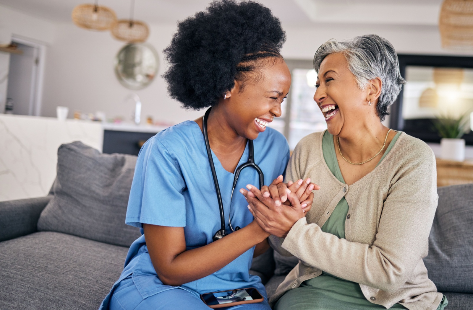 A caregiver and a senior woman in assisted living clasping hands and laughing while sitting on the couch.
