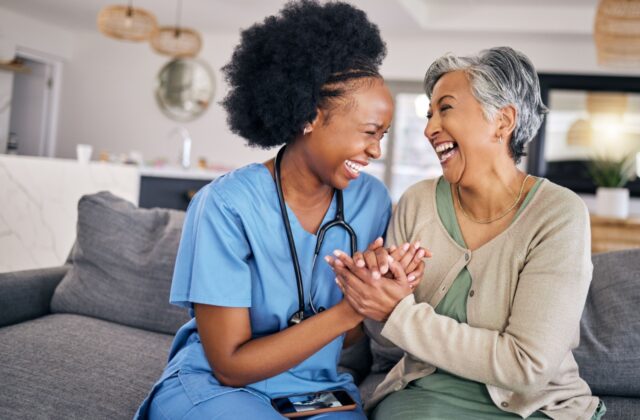 A senior woman and her caregiver in assisted living sitting on the couch, clasping hands, and laughing together.