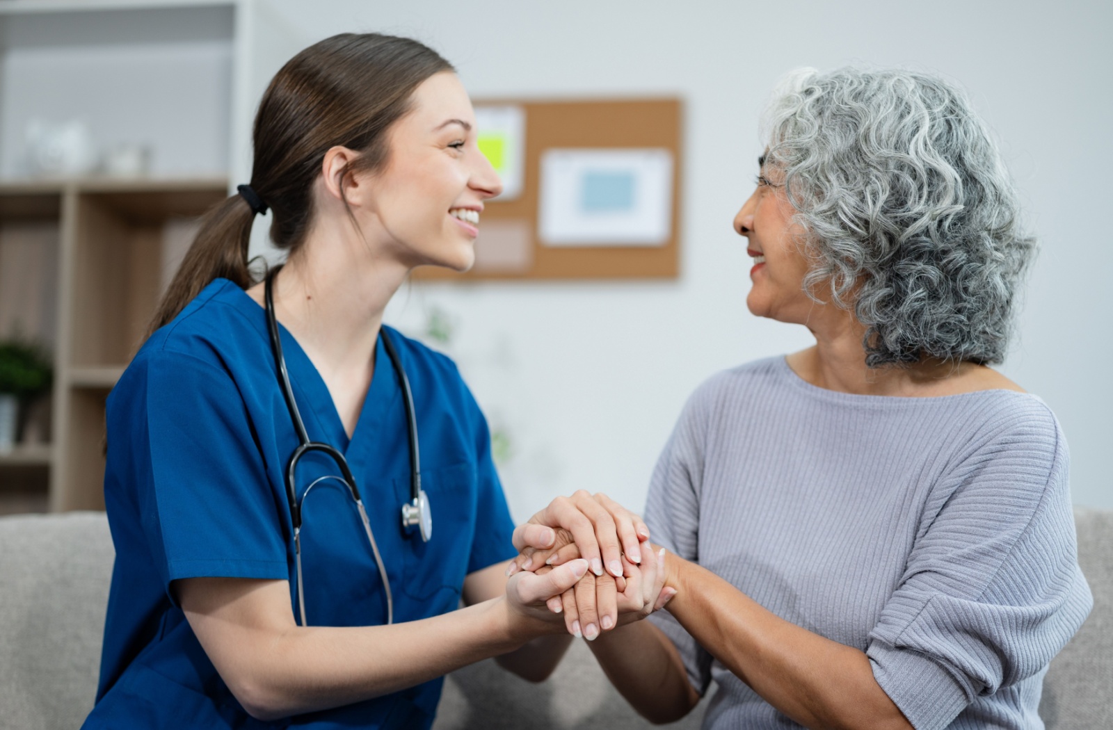 A senior woman in assisted living sitting clasping hands with her caregiver while smiling and sitting on the couch.
