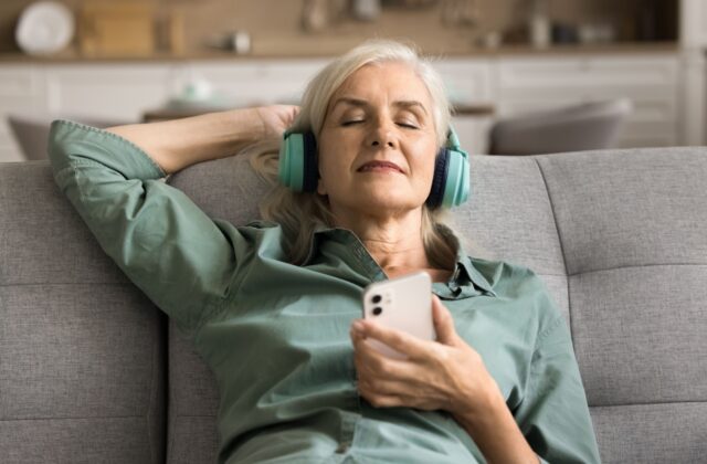 A senior woman relaxing and lying back on the couch, wearing green headphones and listening to an audiobook.