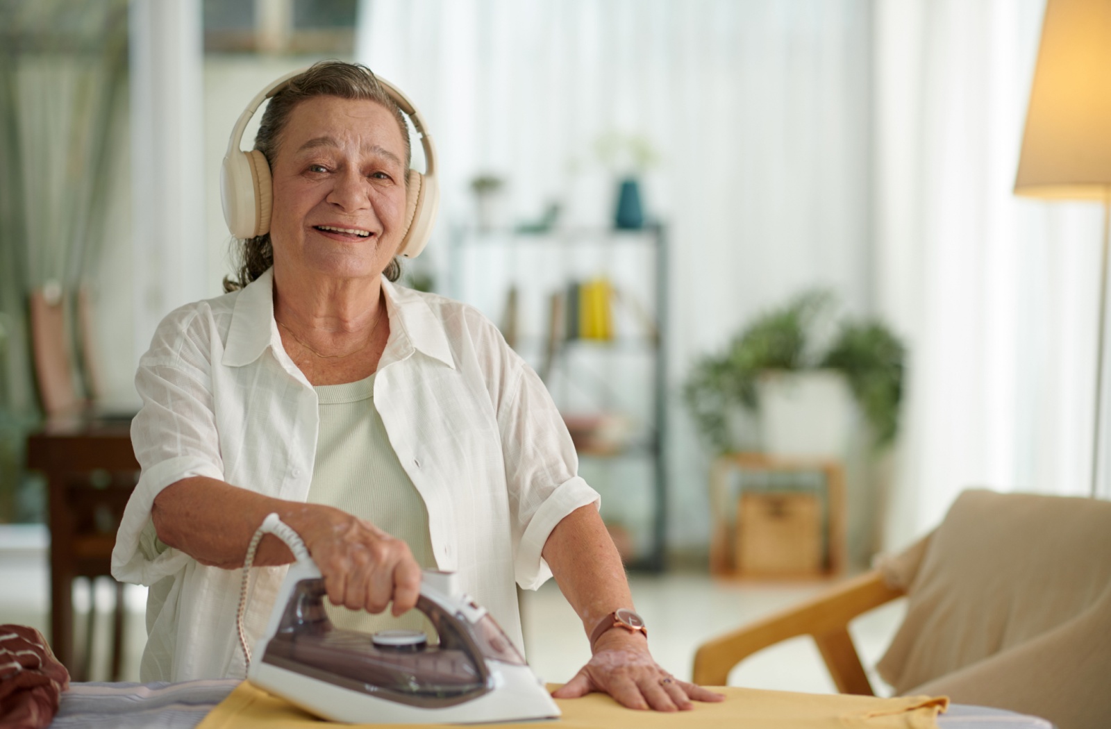 A senior woman smiling, wearing white headphones to listen to an audiobook while ironing a shirt at home.
