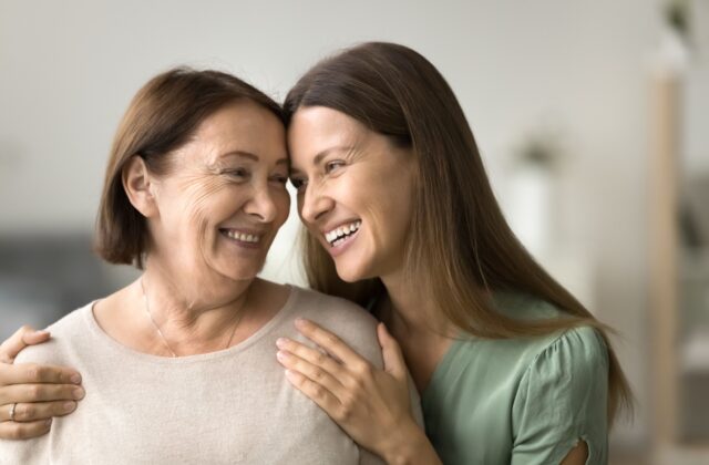 An adult child hugging her senior mom from behind while smiling at one another.