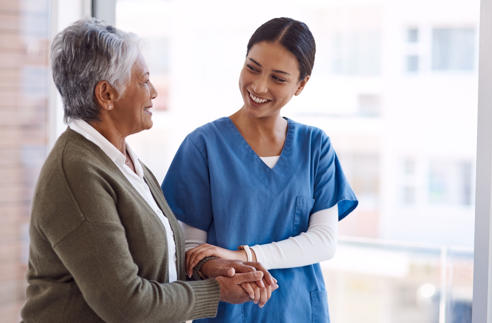 A caregiver and a senior woman walking in front of a window with their arms linked in a senior living community.
