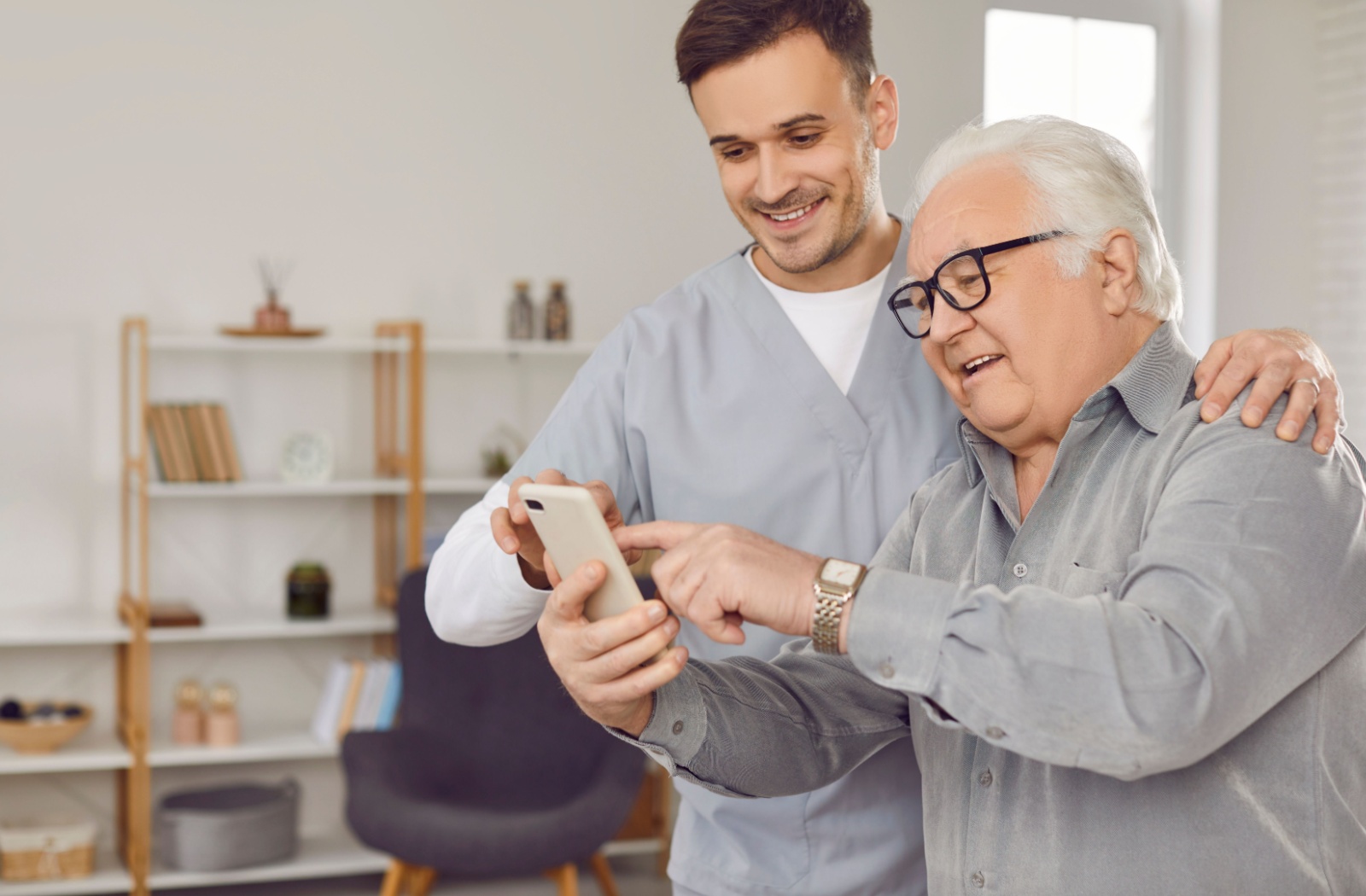 An older man with dementia in memory care smiling while showing off his new cell phone to an excited male caregiver.
